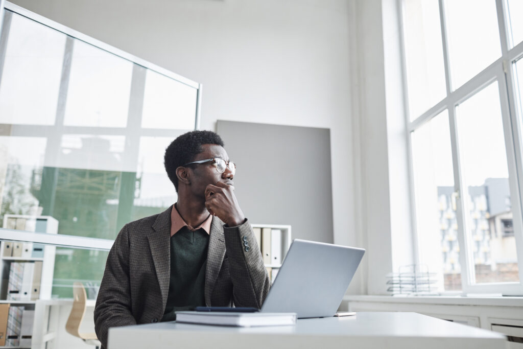 A man sits at a desk with a laptop in an office, looking off into the distance with his hand on his chin while thinking about his career plan.